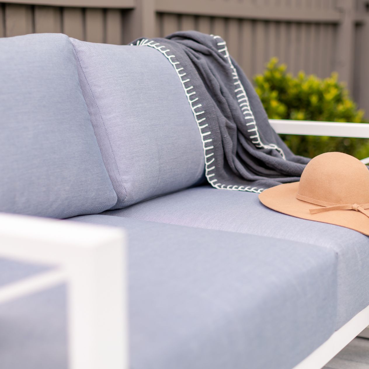 Aluminium outdoor furniture from Linear Lounge collection, featuring a white-framed outdoor lounge with light grey cushions, a brown hat, and plants against a brown fence.