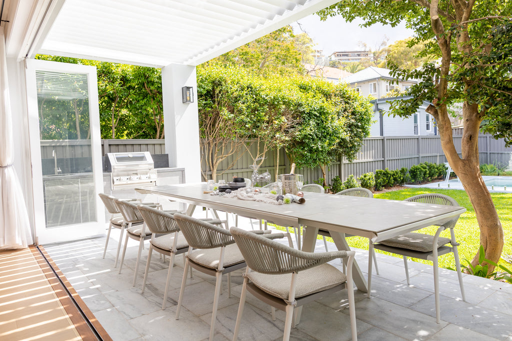 The Neverland Extension Table and Auto Rope Chair 11-Piece Outdoor Dining Setting under a white pergola, with the elegant white rope-woven chairs around the white long table, set against a lush garden and a poolside backdrop.