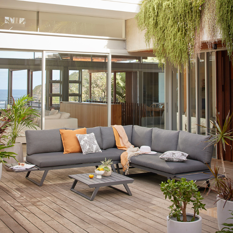 Aluminium outdoor furniture set from Yarra Sofa Series, featuring a 5-seater outdoor lounge with charcoal frame and cushions, teal accent pillows, and a matching coffee table on a wooden deck with a coastal view.